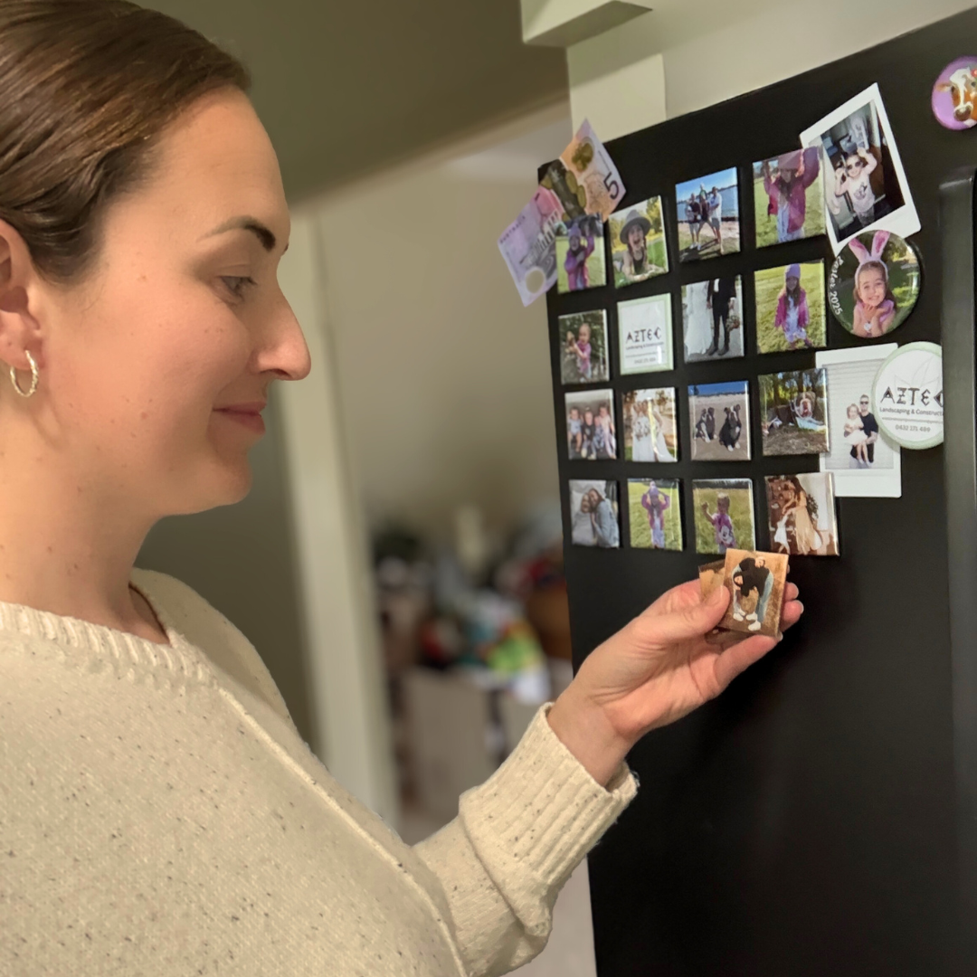 Woman placing handmade photo magnets on a black fridge, showcasing personalised gifts and treasured family memories in an organised display.