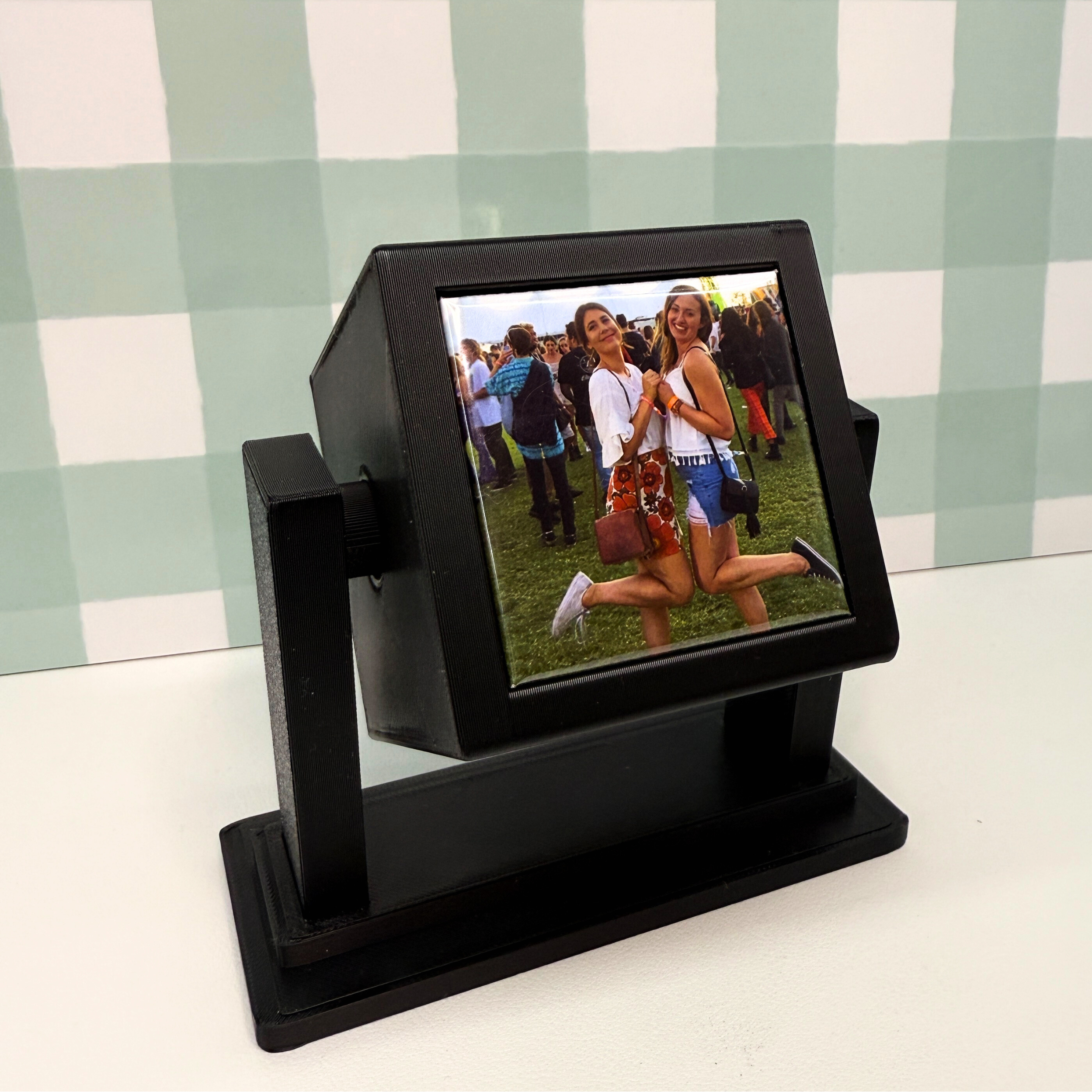 Digital photo frame displaying a photo of two women running on a grassy field with a checkered green and white wall in the background.