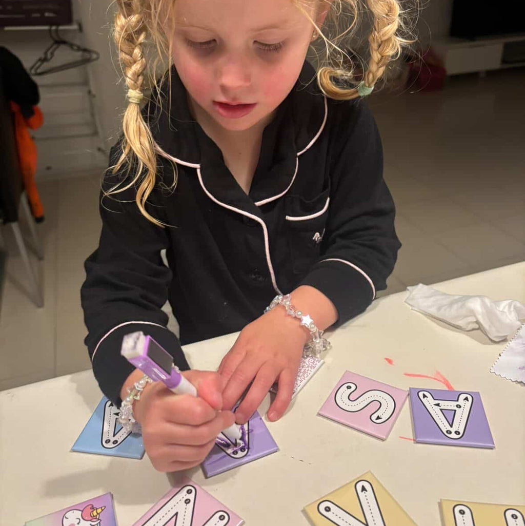 Young girl in black pyjamas practising handwriting by tracing pastel Zara’s World write and wipe magnetic alphabet tiles on a table.