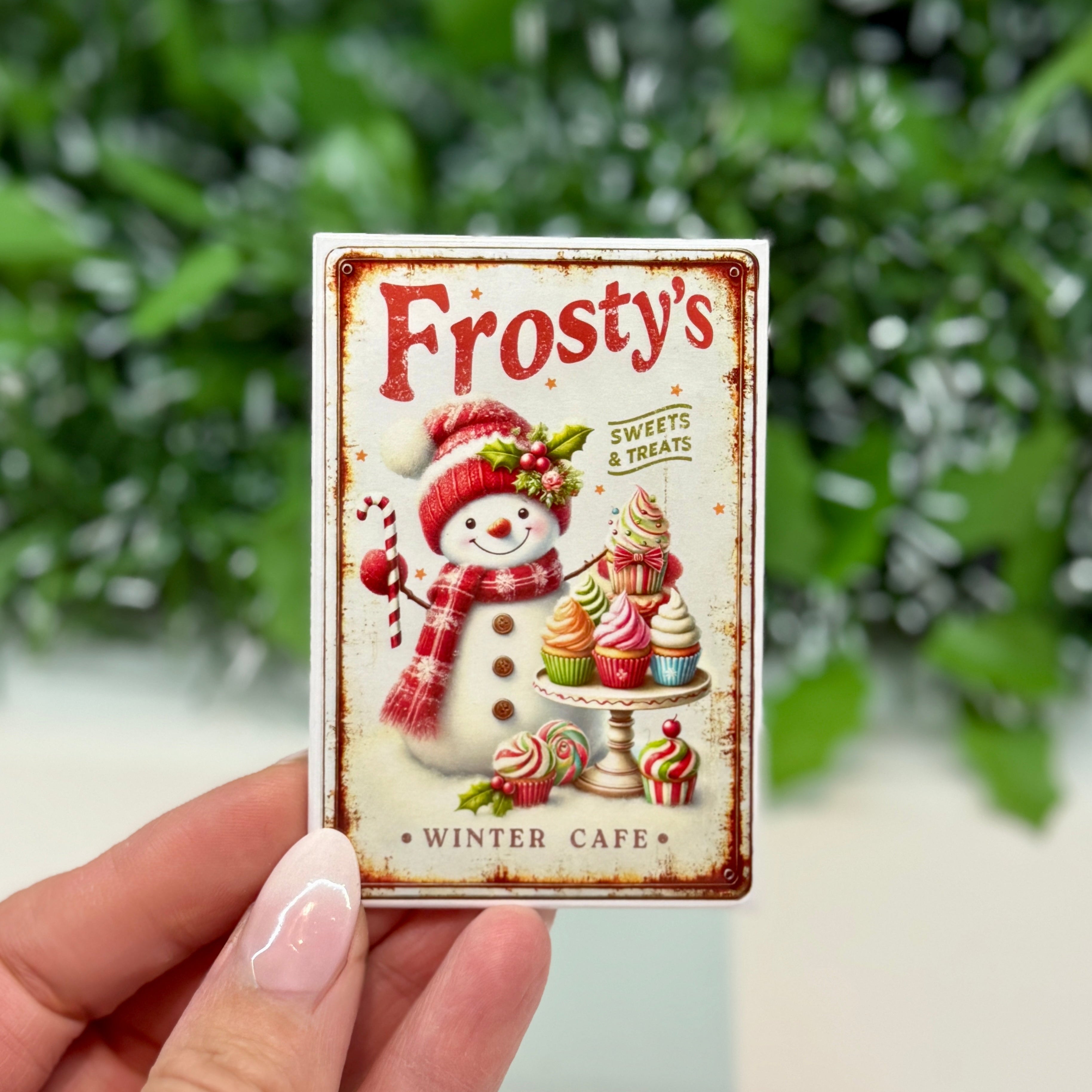 Frosty's Winter Cafe sign held by a hand with a green leafy background