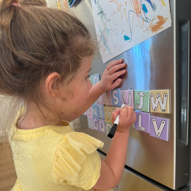 Child in a yellow dress practising letters with Zara’s World pastel write and wipe magnetic alphabet tiles on a fridge.